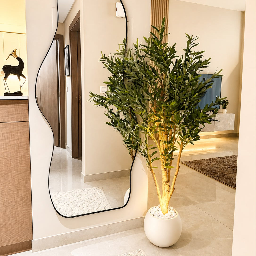 A tall artificial potted plant with green leaves and light brown trunks stands in a white round ceramic pot on a tiled floor in a modern hallway, next to a large wavy-edged floor mirror.